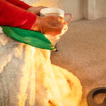 Pensioner trying to keep warm near an electric heater with hot water bottle, blanket and a warm drink. In 2024 the Labour Government in the UK removed the winter fuel allowance from most pensioners.